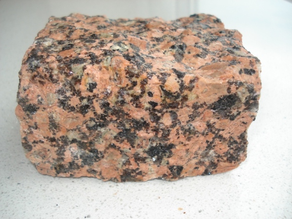 Blok surowego, różowo-szarego granitu z czarnymi plamkami, leżący na jasnej powierzchni.